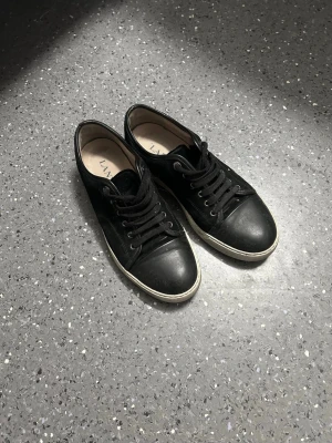 Svarta sneakers från Lanvin - Snygga svarta sneakers från Lanvin med klassisk låg profil och vita sulor. Skorna har svarta skosnören och ovandel i läder som ger en stilren look. Perfekta för dig som gillar minimalistisk design och vill ha något som funkar till det mesta.