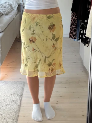 Gul blommig kjol med volangkant - Säljer en ljusgul kjol med blommigt mönster i grönt och brunt. Kjolen har en lätt volangkant nedtill och är tillverkad i ett tunt, flowy material som passar perfekt till varmare dagar. Midjelång modell med normal passform.