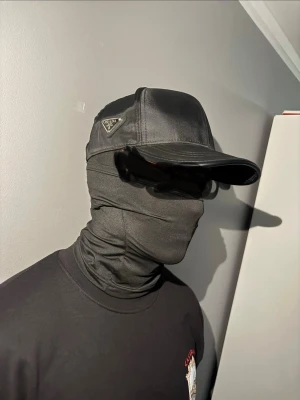 Svart keps från Prada i nylon - Snygg svart keps från Prada i glansigt nylonmaterial med klassisk böjd skärm och diskret logotyp på sidan. Kepsen har en stilren och modern look som passar till streetwear och modeintresserade.