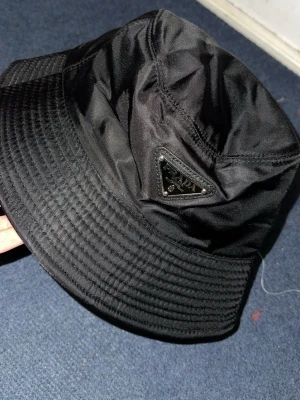 Svart bucket hat från Prada - Snygg svart bucket hat från Prada i glansigt nylonmaterial. Klassisk form med bred, nedåtlutande brätte och ikonisk triangellogo framtill. Perfekt accessoar för en trendig streetwear-look.