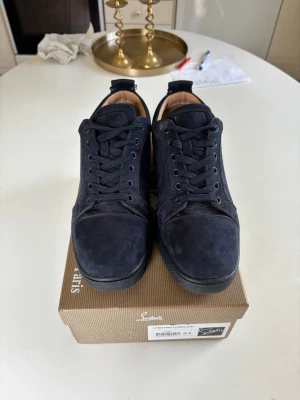 Mörkblå mockasneakers med röd sula - Snygga mörkblå sneakers i mocka från Christian Louboutin med klassisk röd sula och marinblå skosnören . Låg modell med stilren design och exklusiv känsla. Perfekta för dig som vill sticka ut med en ikonisk detalj på fötterna.