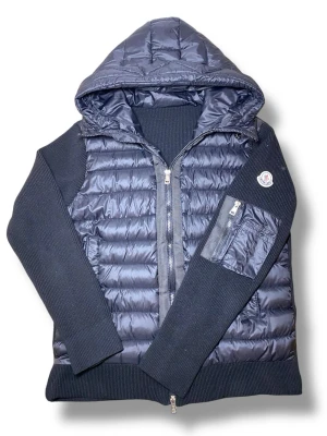 Moncler Cardigan med luva - Snygg svart dunväst från Moncler med quiltad front och rygg, stickade ärmar och sidor samt en praktisk huva. Västen har dragkedja framtill, fickor och en Moncler-logga på ärmen. Perfekt för lager-på-lager och streetstyle.