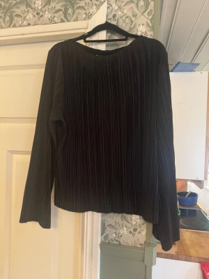 Svart plisserad långärmad topp - Säljer en svart långärmad topp med plisserad struktur. Toppen har en lös passform och rund halsringning, vilket ger en stilren och modern look. Perfekt att matcha med både jeans och kjol för en trendig outfit.