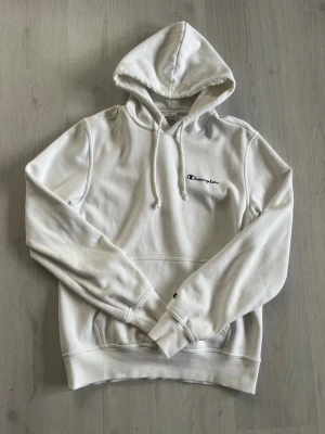 Vit hoodie från Champion - Säljer en stilren vit hoodie från Champion med broderad logga på bröstet. Hoodien har huva med dragsko, magficka och ribbade muddar vid ärmar och nederkant. Perfekt för en avslappnad look.