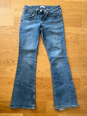 Blå bootcut jeans med fickdetaljer - Säljer ett par klassiska blå bootcut jeans med fem fickor och snygga detaljer på bakfickorna. Jeansen har normal midja och är tillverkade i ett mjukt denimtyg med lätt tvättad look. Perfekta för en avslappnad och trendig stil.