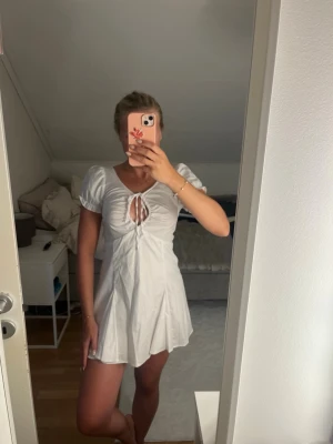Vit kort klänning med snörning - Säljer en söt vit kortklänning med puffärmar och snörning framtill. Den har shorts under och är endast använd 1 gång så väldigt bra skick. 