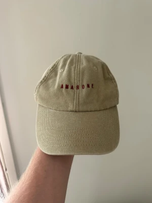 Cocktailored Keps Amarone Beige  - Snygg beige keps från Cocktailored med böjd skärm och broderad text 'AMARONE' i rött framtill. Klassisk sexpanel-design med ventilationshål och justerbar rem bak. Tillverkad i bomullstyg för en avslappnad och stilren look.