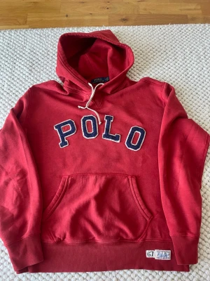 Röd hoodie från Polo Ralph Lauren - Röd hoodie från Polo Ralph Lauren med stor POLO-text i blått och vitt på bröstet. Klassisk känguruficka framtill, ribbade muddar och nederkant samt vit dragsko i huvan. Märkeslapp nertill och mjukt bomullsmaterial. Perfekt för en avslappnad och sportig stil.