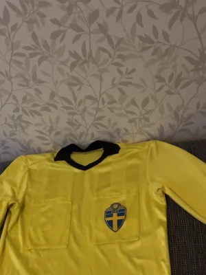 Gul långärmad fotbollströja Sverige - Klassisk gul långärmad fotbollströja med svart krage och Sveriges landslagsemblem på bröstet. Tröjan har två bröstfickor och är tillverkad i ett glansigt syntetmaterial. Perfekt för dig som vill sticka ut på planen.