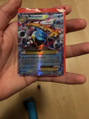 Pokémonkort Mega Blastoise EX - Säljer ett Pokémonkort av typen Mega Blastoise EX med 220 HP. Kortet har en färgstark och glansig design med blå, röd och gul färg. Det är inplastat i ett rött plastfodral och har attacken Hydro Bombard. Perfekt för samlare eller Pokémonfans.