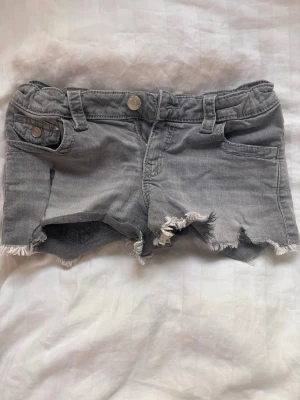 Grå jeansshorts med fransig kant - Snygga grå jeansshorts med fransig och rå kant nertill. Klassisk femficksmodell med knapp och dragkedja framtill samt bälteshällor. Bakfickor med lock och knappdetalj. Perfekta för varma dagar.