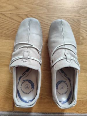 Beige loafers med vita detaljer - Snygga beige loafers i mockaimitation med vita dekorativa band över foten. Skorna har en fyrkantig tå och synliga sömmar framtill för en modern look. Perfekta för dig som vill ha en stilren och bekväm sko till vardagsoutfiten.