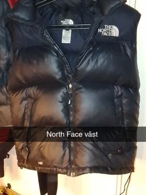 Svart dunväst från The North Face - Snygg svart dunväst från The North Face med dragkedja framtill och hög krage. Västen har logga på bröstet och är riktigt puffig för extra värme. Perfekt för lager på lager och streetwear-stil.
