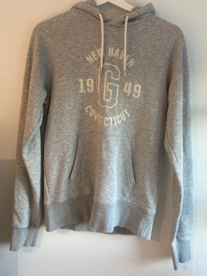 Grå GANT hoodie - Snygg grå hoodie med huva och vita dragsnören. Tryck på bröstet med text och känguruficka framtill. Skön hoodie som är både lång och luftig.