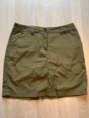 Olivgrön skort från Jack Wolfskin - Snygg olivgrön skort från Jack Wolfskin med klassisk look. Kjol med inbyggda shorts, två sidofickor och bälteshällor. Tillverkad i slitstarkt bomullsmaterial, perfekt för dig som gillar outdoor och vill ha både stil och funktion.