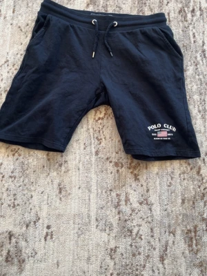 Mörkblå shorts med snörning Polo Club - Mörkblå shorts från Polo Club med snörning i midjan och tryckt logga på vänster ben. Mjukt material och en bakficka. Perfekta för sommaren och chill dagar.