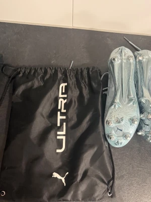 Puma Ultra fotbollsskor ljusblå - Säljer ett par Puma Ultra fotbollsskor i en snygg ljusblå färg. Skorna har metallskruvdobbar och är tillverkade i ett lätt syntetmaterial för maxad speed på planen. Medföljer gör en svart Ultra-påse med Puma-logga. Perfekta för dig som vill dominera på gräs. Modell är elite så att säga. Pris ej hugget i sten. De är elit och knappt använda satte på mej skorna och kände de va lite för små så mer eller mindre helt nya