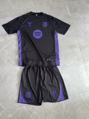 Svart och lila FC Barcelona träningsset - Säljer ett svart och lila träningsset med FC Barcelona-logga och Kobe Bryant-symbol på både tröja och shorts. Setet har lila detaljer längs sidorna och är gjort i ett lätt och ventilerande syntetmaterial, perfekt för träning eller fotboll.