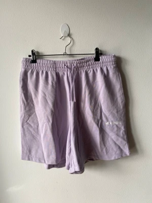 Ljuslila shorts från McKenzie - Säljer ett par ljuslila shorts från McKenzie med elastisk midja och snörning. Shortsen har en avslappnad passform och är tillverkade i mjuk bomull, perfekt för varma dagar. Diskret logga på vänster ben för en clean look.