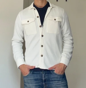 Vit overshirt - Vit långärmad overshirt med två bröstfickor och bruna knappar. Skjortan har en klassisk krage och är tillverkad i ett strukturerat bomullstyg som ger en avslappnad men snygg look. Helt oanvänd.
