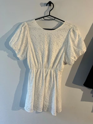 Vit kort klänning med broderi och puffärm - Söt vit kortklänning med broderade blommor, puffärmar och elastisk midja. Klänningen har rund halsringning fram och öppen rygg med knytband. Perfekt för sommarens alla tillfällen!