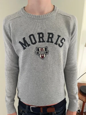 Grå tröja från Morris med logga - Snygg grå stickad tröja från Morris med svart text och broderad logga på bröstet. Tröjan har rund halsringning, långa ärmar och ribbade muddar. Perfekt för en stilren och avslappnad look.                                                                   Skickas inom 1-3 dar vid beställning 