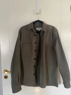 Olivgrön overshirt från Just Junkies - Säljer en olivgrön overshirt från Just Junkies i storlek S. Skjortan har två bröstfickor, svarta knappar och klassisk krage. Materialet är kraftigt bomullstyg som ger en avslappnad men stilren look. Perfekt att bära öppen över en t-shirt eller skjorta.