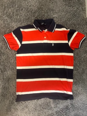 Randig pikétröja från Polo Ralph Lauren - Säljer en snygg randig pikétröja från Polo Ralph Lauren i rött, marinblått och vitt. Tröjan har klassisk krage, knappar vid halsen och den ikoniska broderade loggan på bröstet. Perfekt till jeans eller shorts för en chill look.