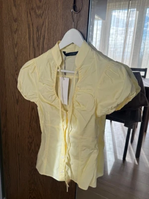 Poplintopp Zara - Söt ljusgul blus med puffiga korta ärmar och krage. Blusen har knappar framtill och en dekorativ knytning vid halsen. Tillverkad i ett tunt, lätt material. Perfekt för dig som gillar en feminin och trendig stil.