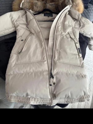 Beige dunjacka med fuskpäls Cedrico - Snygg beige dunjacka från Cedrico i storlek XS. Jackan har två stora framfickor med dragkedja och en huva med mjuk fuskpäls i bruna toner. Perfekt för kalla vinterdagar och har en längre modell för extra värme.