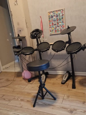 Elektroniskt trumset med pall - Säljer ett komplett elektroniskt trumset från Alesis med flera pads, två pedaler och tillhörande trumstol. Setet är svart och har ett stativ i metall samt hörlurar. Perfekt för dig som vill öva trummor hemma utan att störa grannarna.tillhör ochså trummpinnar och allt är i ny skick