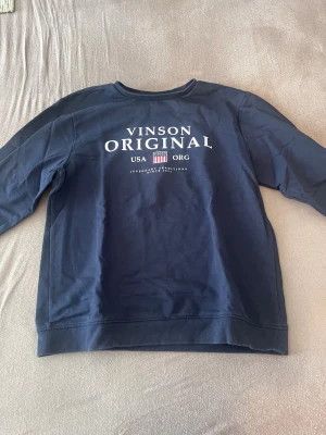 Mörkblå sweatshirt Vinson Original - Mörkblå sweatshirt från Vinson Original med tryck framtill i vitt och rött. Tröjan har rund halsringning och långa ärmar. Tillverkad i mjuk bomull för en bekväm känsla. Perfekt för en avslappnad och stilren look.