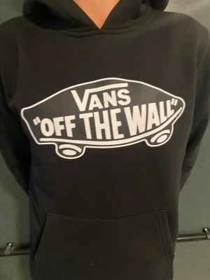 Svart Vans hoodie med tryck - Säljer en svart hoodie från Vans med det klassiska 'Off The Wall'-trycket i vitt på bröstet. Hoodien har huva och en stor magficka framtill. Perfekt för dig som gillar streetwear och vill ha en skön och stilren tröja.