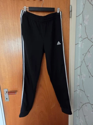 Svarta Adidas mjukisbyxor med ränder - Svarta mjukisbyxor från Adidas med vita ränder längs sidorna och Adidas-logga på ena benet. Byxorna har resår i midjan och är tillverkade i ett mjukt och stretchigt material. Perfekta för en avslappnad och sportig stil. Slitna över hela byxorna (se bild 4)