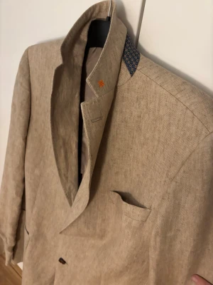 Beige linnékavaj från Morris - Stilren beige kavaj från Morris i linnematerial. Kavajen har två knappar framtill, bröstficka och klassisk krage med en liten orange detalj. Insidan är fodrad i beige med lila kantband och flera innerfickor. Perfekt för en dressad look. Nypris 2900