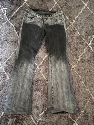 Grå bootcut jeans med tvättad look - Säljer ett par grå bootcut jeans med tydlig tvättad effekt och slitningar. Jeansen har låg midja, fem fickor och vida ben. Materialet är klassisk denim i bomull. Perfekta för dig som gillar retrostil och statement-plagg.