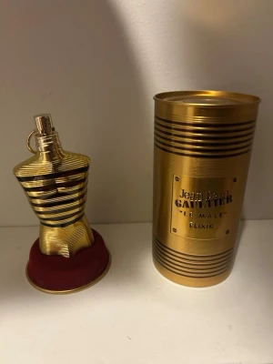 Jean Paul Gaultier Le Male Elixir - Exklusiv parfym i en guldig flaska formad som en manlig torso med svarta ränder. Kommer med en matchande guldfärgad metallburk och röd sammetsbas. Flaskan har en unik och lyxig design som verkligen sticker ut i badrumshyllan.