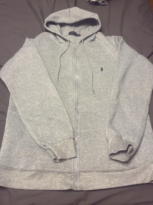 Grå zip hoodie från Polo Ralph Lauren - Snygg grå hoodie med dragkedja från Polo Ralph Lauren. Klassisk design med huva och dragsnören, samt den ikoniska loggan broderad på bröstet. Tillverkad i mjukt sweatshirtmaterial som är perfekt för chill dagar. Passar till allt och är superbekväm.