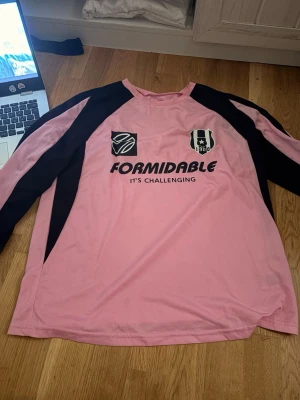 Rosa långärmad sporttröja - Säljer en rosa långärmad sporttröja med svarta detaljer längs ärmar och axlar. Tryck på bröstet med texten 'FORMIDABLE IT'S CHALLENGING' samt två loggor, varav en med årtalet 1908. Perfekt för träning eller match.