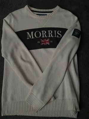  Morris Tröja | S - Vit stickad tröja från Morris med svart panel och stor logga framtill samt brittisk flagga. Tröjan har rund halsringning, ribbade muddar och patch på ärmen. Perfekt för dig som gillar klassisk och stilren design.