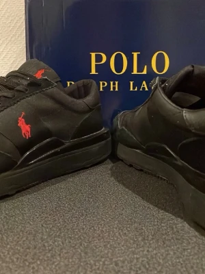 Polo Ralph Lauren skor | 41 - Snygga svarta sneakers från Polo Ralph Lauren med klassisk röd logga på sidan. Skorna har en låg profil, svarta snören och en chunky sula. Materialet ser ut att vara en mix av mocka och textil för en modern look.