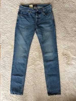 Neuw Stockholm jeans  - Säljer ett par blå jeans med smal passform och ett unikt svartvitt tryck på ena bakfickan. Jeansen har klassisk femficksdesign, knappgylf och är tillverkade i denim. Perfekta för dig som vill ha något som sticker ut lite extra.