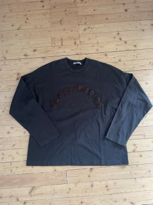 Acne Studios sleeve  - Säljer en mörkgrå långärmad topp från Acne Studios med broderad logga i brunt framtill. Oversized passform och rund halsringning. Perfekt för dig som gillar streetwear och vill ha något stilrent men ändå unikt.