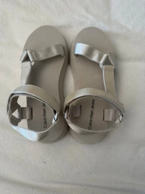 Beige sandaler från Calvin Klein Jeans - Snygga beige sandaler från Calvin Klein Jeans med breda remmar och justerbart spänne runt ankeln. Sandalerna har en chunky platåsula och är tillverkade i syntetmaterial med textilband. Perfekta för en trendig och avslappnad look.