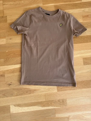 Beige t-shirt från lyle&scott - Snygg beige t-shirt i bomull med korta ärmar och rund hals. På bröstet sitter en liten broderad logga i gult och svart som ger en clean och stilren look. Perfekt till jeans eller shorts för en enkel outfit.