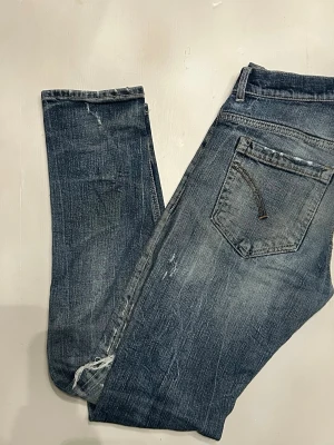 Dondup - Säljer ett par blå jeans med slitningar och hål på både fram- och baksida. Jeansen har klassisk femficksmodell, normal midja och raka ben. Materialet är denim i bomull och de har en cool, avslappnad look med råa kanter och distressed detaljer. 