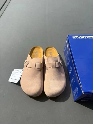  Birkenstock Boston Mud - Säljer ett par snygga beige tofflor från Birkenstock i mjuk mocka. Modellen har en klassisk slip-in design med justerbart spänne och ergonomisk fotbädd för extra komfort. Perfekta för chill dagar hemma eller när du vill ha en avslappnad stil.
