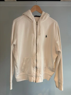 Vit hoodie med dragkedja från Polo Ralph Lauren - Vit hoodie från Polo Ralph Lauren med dragkedja framtill, huva med snörning och två fickor. Klassisk broderad logga på bröstet. Perfekt för en chill och stilren look.Nypris ligger på 2200 kr. Personen på bilden är 175 cm. 