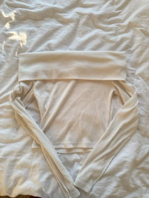 Offshoulder ribbad topp i vitt - Snygg offshoulder topp i vitt med ribbad struktur och långa ärmar. Perfekt för dig som vill ha en stilren och trendig look. Toppen är figurnära och har en bred kant upptill som ger en cool touch.