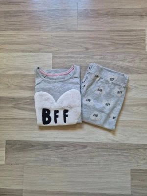 Grå pyjamas med BFF-hjärta - Mysig grå pyjamas med långärmad tröja och byxor. Tröjan har ett stort vitt hjärta i fluffigt material med texten 'BFF' på bröstet. Byxorna har tryck med små 'BFF'-texter över hela. Perfekt för sleepovers eller hemmamys.